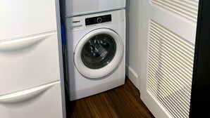 A compact laundry room with a stacked washer and dryer, a wooden floor, and a white door with blinds. - Downtown LA - Free Parking- Legends Loft! (Vernon)