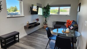 Een moderne woonkamer met een ronde glazen tafel, zwarte stoelen, een donkere bank, een wandtelevisie en een potplant.