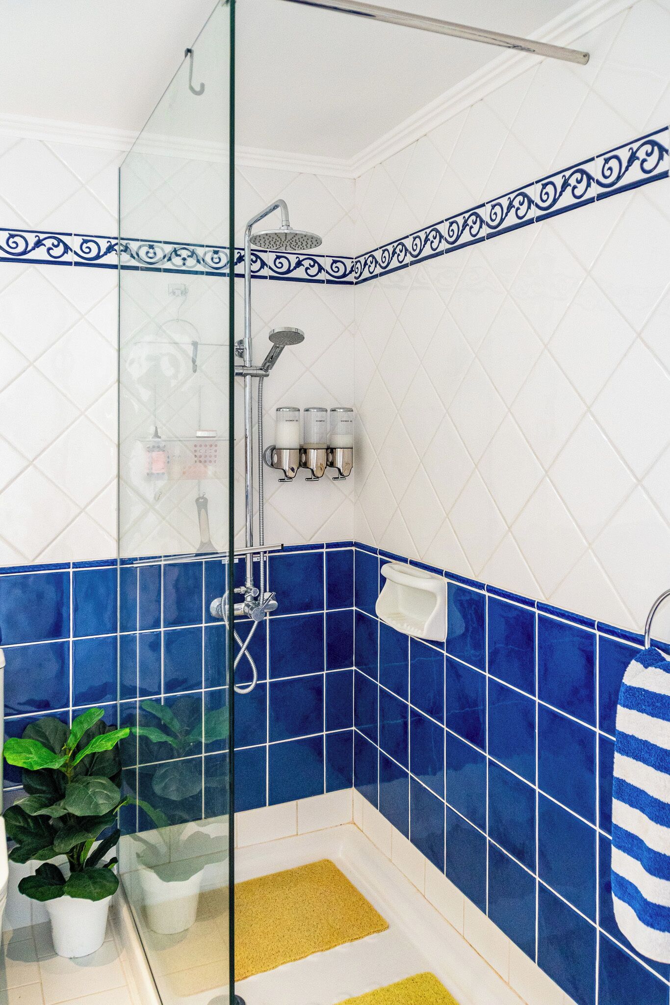 Ein Badezimmer mit blau-weißen Fliesen, einer Duschkabine aus Glas, einem Blumentopf und einer gelben Badmatte.