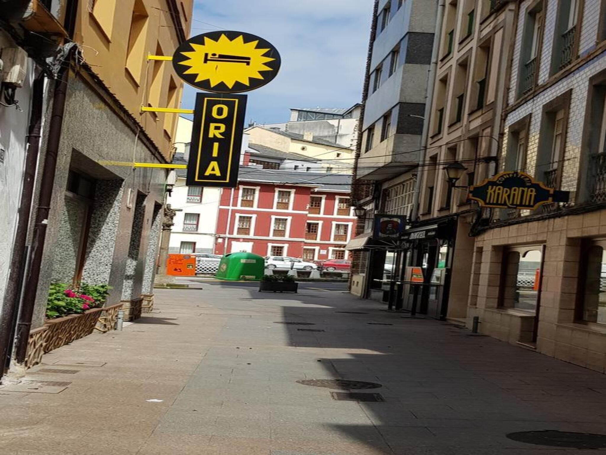 A narrow street with buildings on either side, a sign for 'ORIA' above a yellow awning, and a 'KARANA' sign on the right.