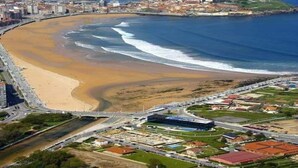 Vista aerea di una zona costiera con una spiaggia sabbiosa curvilinea, un porto turistico e un mix di edifici residenziali e commerciali.