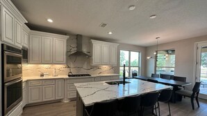 A modern kitchen with white cabinetry, a marble island, and a dining area with a large table and chairs.