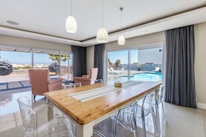 A modern dining area with a wooden table, clear chairs, and pendant lights. It features large windows with a view of a pool and outdoor seating. - Beachfront, Private Pool, Pizza Oven, Pool Table, Fast Internet, Family Friendly (Cape Town)