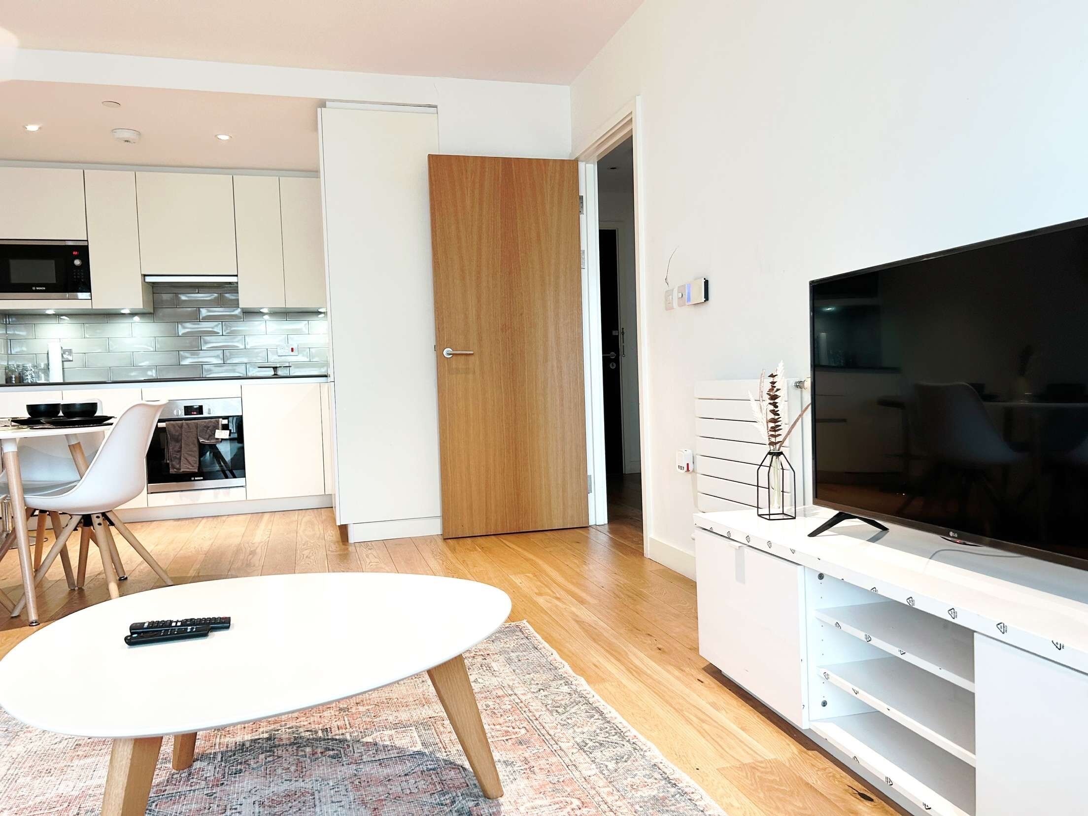 Et moderne køkken med hvide skabe, en mikrobølgeovn og ovn. Der er et spiseområde med et hvidt bord og stole, samt et fjernsyn på et stativ med hylder.