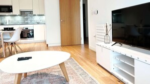A modern kitchen with white cabinets, a microwave, and an oven. There's a dining area with a white table and chairs, and a TV on a stand with shelves below. - Stylish Apt Sleeps 2 with Balcony (London)