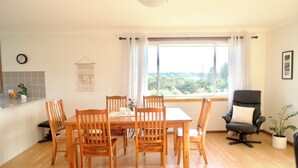 A dining area with a wooden table and chairs, a black office chair, a window with a view of greenery, and a wall-mounted clock.