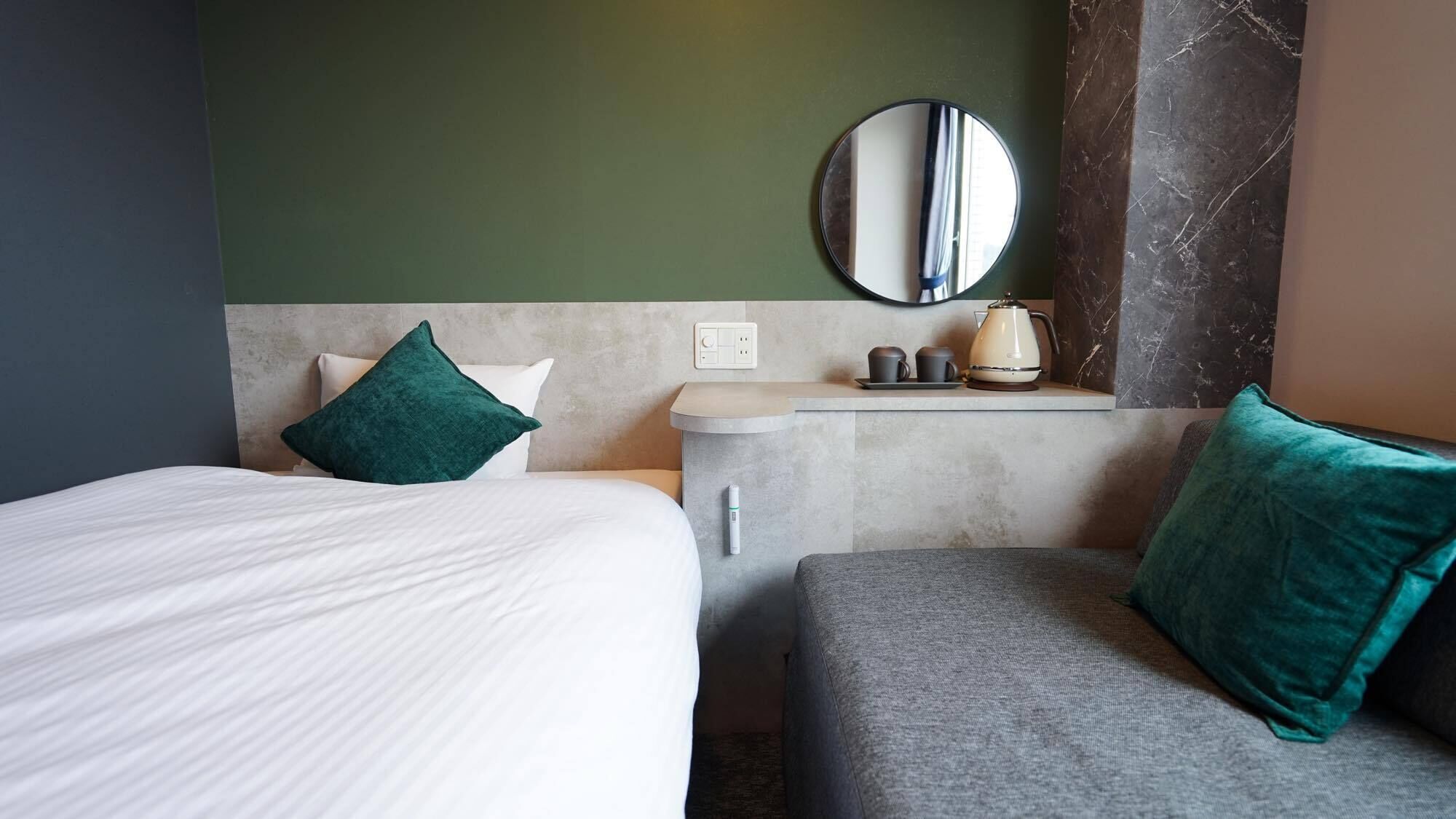 A hotel room with a bed, a sofa, a small table with a kettle, a mirror, and a wall socket.