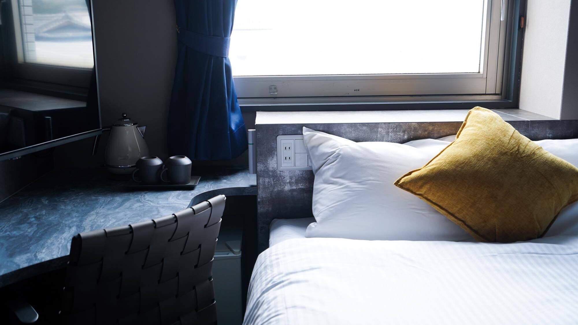 A hotel room with a bed, a desk with a kettle, and a chair.
