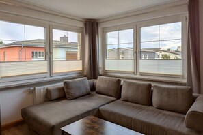 A modern living room with a sectional sofa, a small wooden table, and large windows offering views of neighboring buildings. - Your perfect vacation home: Cozy vacation apartment Franke 1.2 in Zinnowitz! - (Zinnowitz)