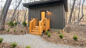 Una petita cabana de fusta amb una escala de fusta que mena a una porta, envoltada d'arbres i un camí de grava.