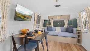Interior d'una caravana domotècnica amb zona de menjador, sofà i televisió que mostra un paisatge idíl·lic.