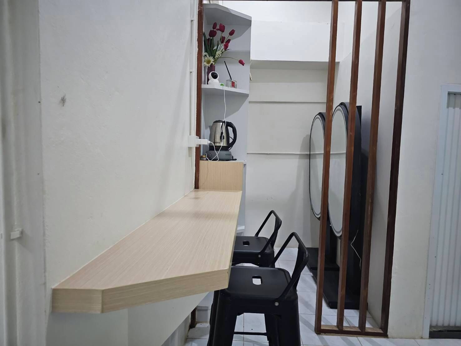 A small, compact room with a wooden desk, two black chairs, a white shelf with flowers, a kettle, and a mirror.