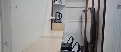 A small, compact room with a wooden desk, two black chairs, a white shelf with flowers, a kettle, and a mirror.