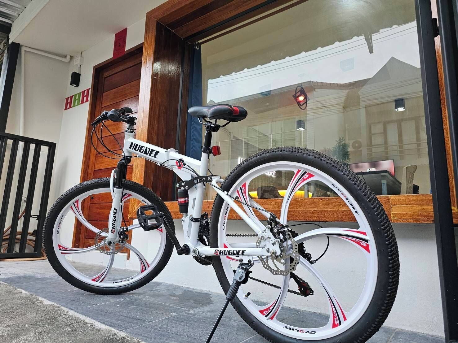 Un vélo de montagne blanc, orné de la marque « HUGDEE », est garé devant un bâtiment doté d’une porte en bois et d’une fenêtre en verre qui reflète un autre bâtiment.