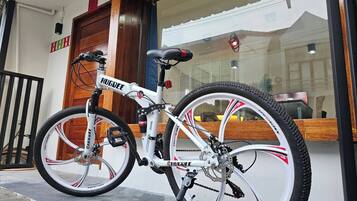 Un vélo de montagne blanc, orné de la marque « HUGDEE », est garé devant un bâtiment doté d’une porte en bois et d’une fenêtre en verre qui reflète un autre bâtiment.