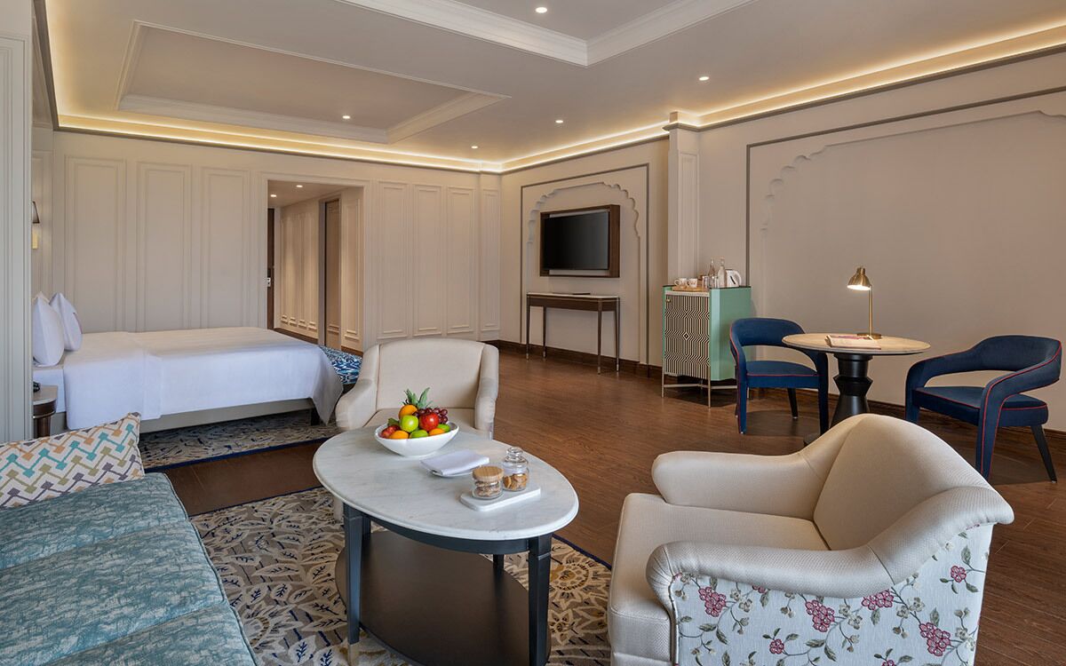 Una camera d'albergo con un letto, un divano, un tavolino con una ciotola di frutta, due poltrone, una televisione e un piccolo tavolo con una lampada.