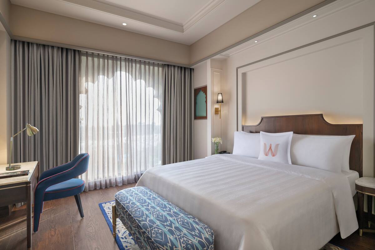 Een hotelkamer met een groot bed, een bureau, een stoel en uitzicht op het groen door dunne gordijnen.