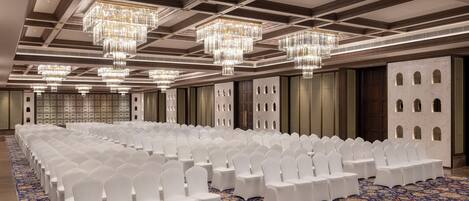 Un amplio salón de conferencias con hileras de sillas blancas, candelabros y una alfombra estampada.