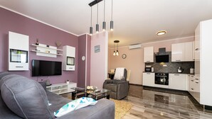 Ein modernes Wohnzimmer mit grauem Sofa, Flachbildfernseher und Glastisch. Die Küche verfügt über weiße Schränke, ein schwarzes Backrohr und eine Mikrowelle.