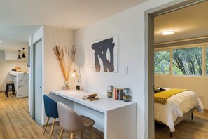 A modern bedroom with a bed, a desk with a chair, a painting, and a window with a view of trees.