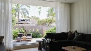 Una sala de estar moderna con un sofá oscuro, una mesita redonda y vistas a un jardín trasero con plantas y una zona de patio.
