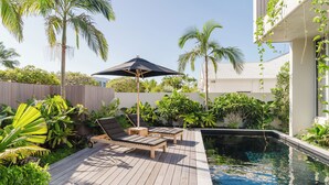 Una terraza de madera con dos sillones de descanso y una mesa, una piscina y exuberante vegetación tropical.
