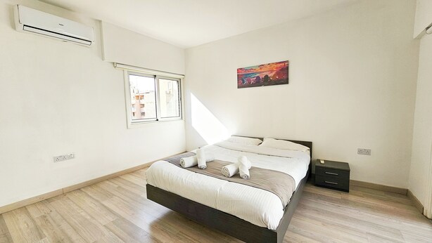 A bedroom with a bed, a nightstand, an air conditioning unit, a window, and a painting on the wall. - Blue Sky Ocean Retreat (Larnaca)