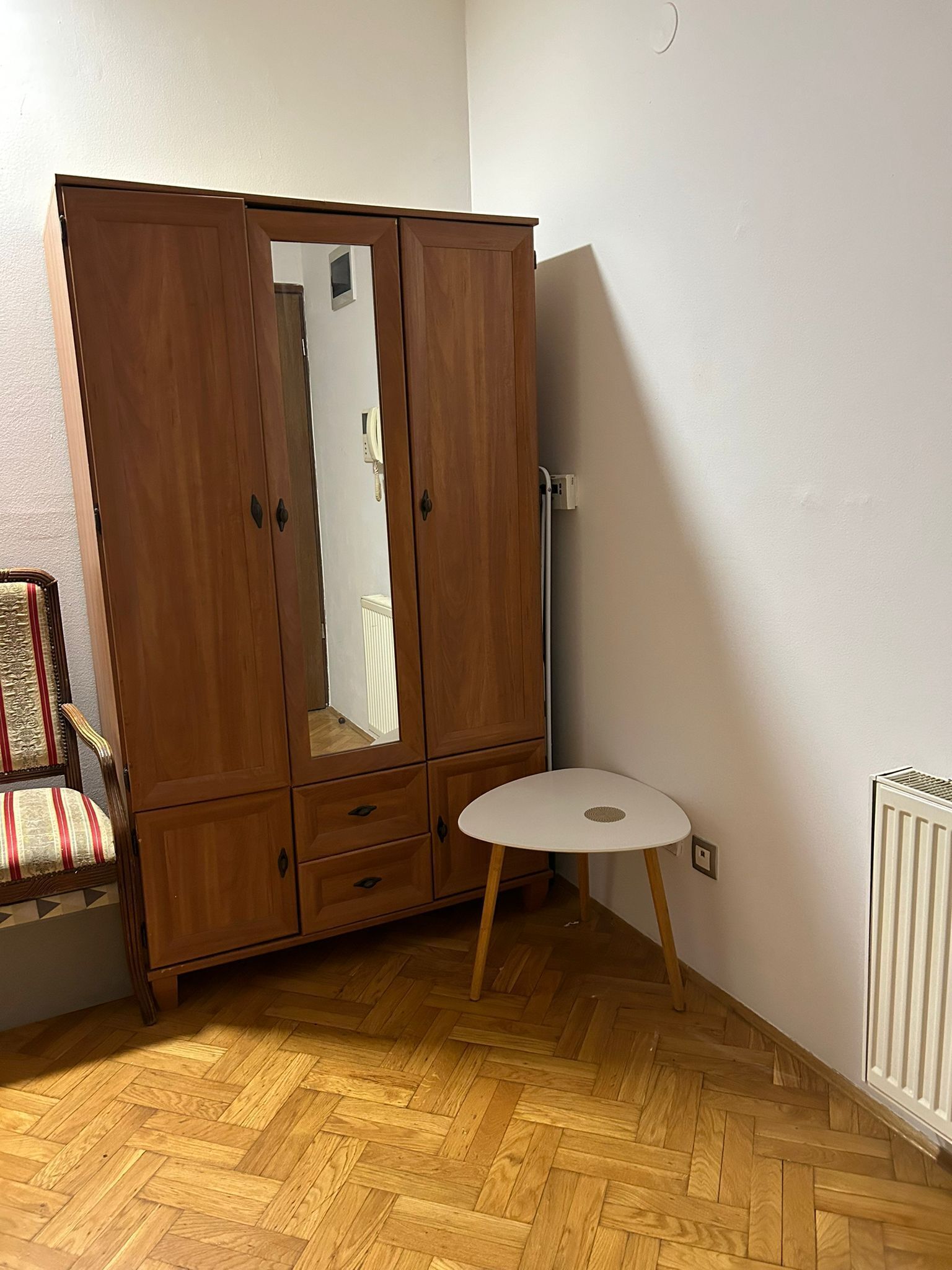 A room with a wooden wardrobe, a small round table, a chair with a striped cushion, and a radiator.