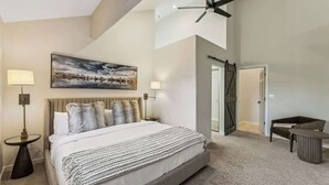 A bedroom with a large bed, two bedside lamps, a ceiling fan, a painting, and a door leading to another room.