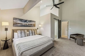 Une chambre à coucher avec un grand lit, deux lampes de chevet, un ventilateur de plafond, un tableau et une porte donnant sur une autre pièce.