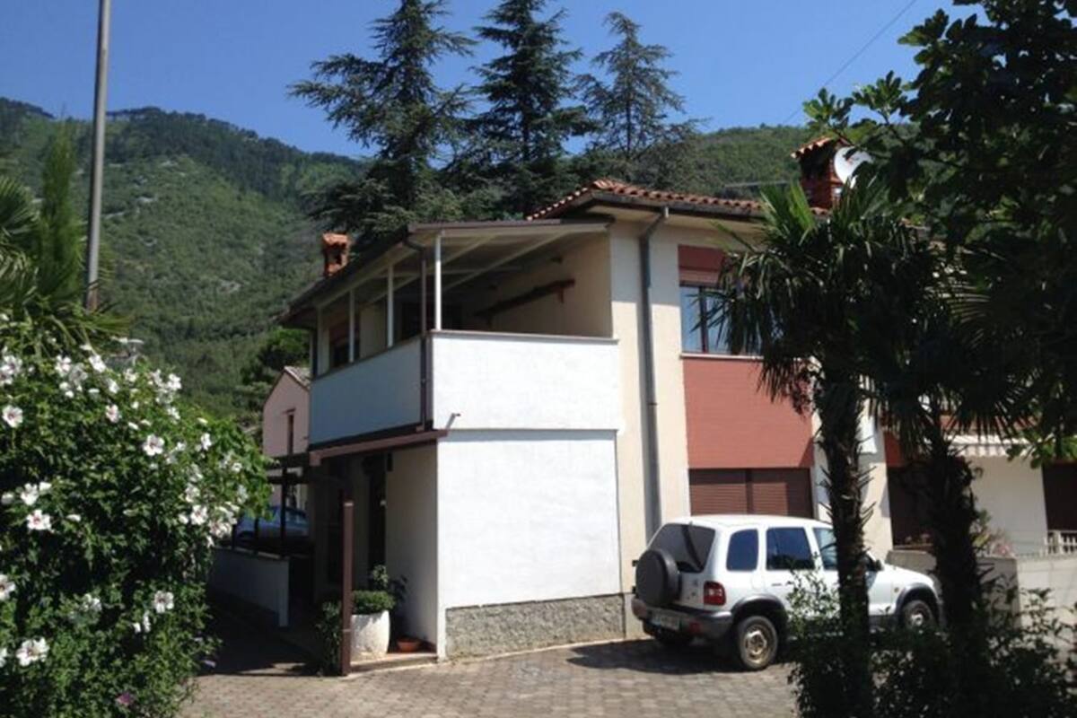 Ein zweistöckiges Haus mit Balkon, einem parkenden SUV und einer Bergkulisse im Hintergrund.