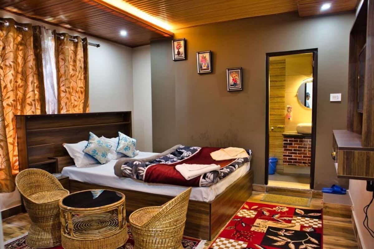 A bedroom with a bed, two wicker chairs, a wooden headboard, a red and black patterned rug, and a small bathroom visible through an open door.