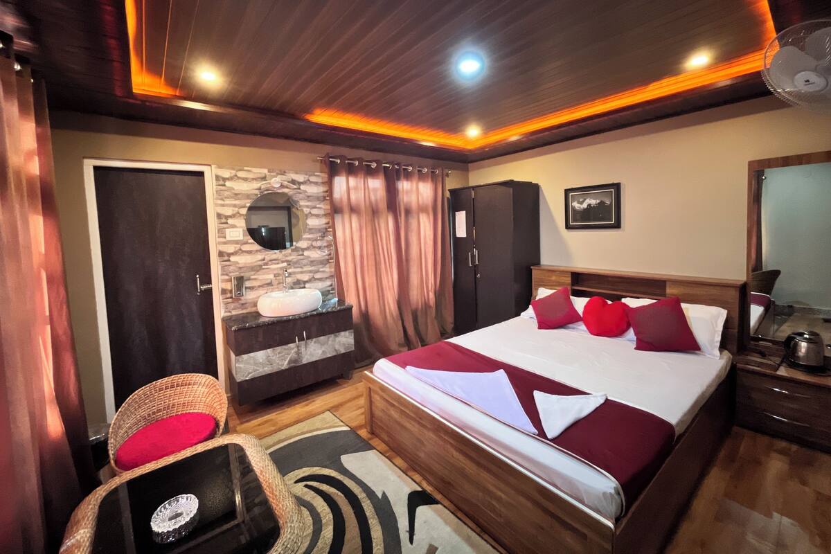 A bedroom with a bed, a wooden wardrobe, a bathroom with a sink and mirror, and a wicker chair.
