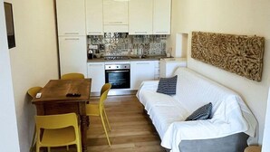 A compact living space with a kitchenette, a dining area with a wooden table and yellow chairs, and a sofa with patterned cushions. - La Perla del Cavo, just a stone's throw from the splendid seafront! (Cavo)