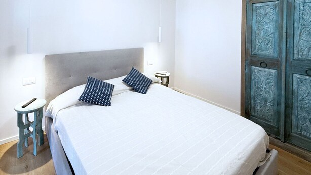 A neatly made bed with a grey headboard, two bedside tables with lamps, and a blue door with intricate designs. - La Perla del Cavo, just a stone's throw from the splendid seafront! (Cavo)