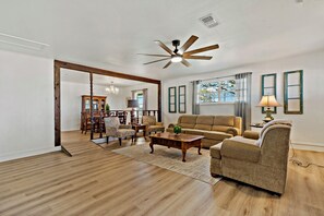A spacious living room with a wooden floor, a ceiling fan, a sofa, a coffee table, and a dining area with chairs and a chandelier.