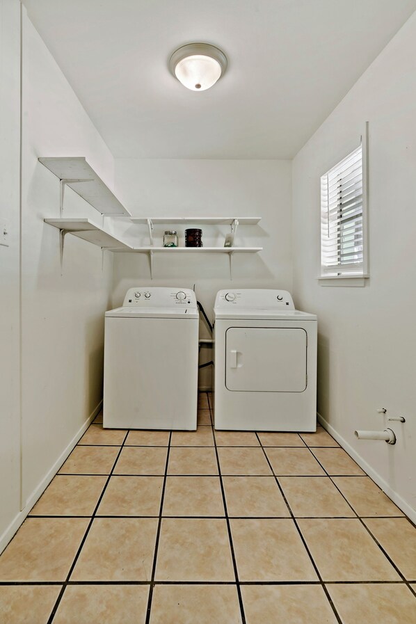 A laundry room with two white washing machines, a shelf above, and a window with blinds.