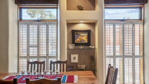 A dining area with a wooden table, chairs, and a red and blue woven placemat. There are two windows with white shutters, a framed painting, and a decorative bowl on a shelf. - Casa Feliz I Elegant 2-Bedroom Santa Fe Retreat (Santa Fe)