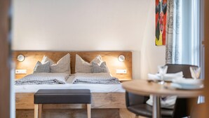 Ein Bett mit Holz-Kopfteil, zwei Nachttische, eine Bank und ein runder Tisch mit Stühlen.