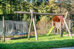 A playground with swings and a slide, surrounded by a wooden fence and a house with a red roof.