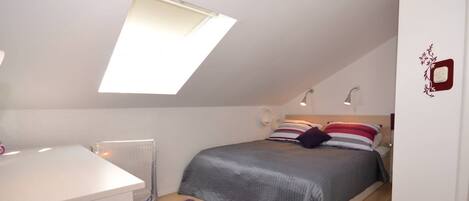 A small, well-lit bedroom with a sloped ceiling, a bed with striped bedding, a purple rug, and a white desk.