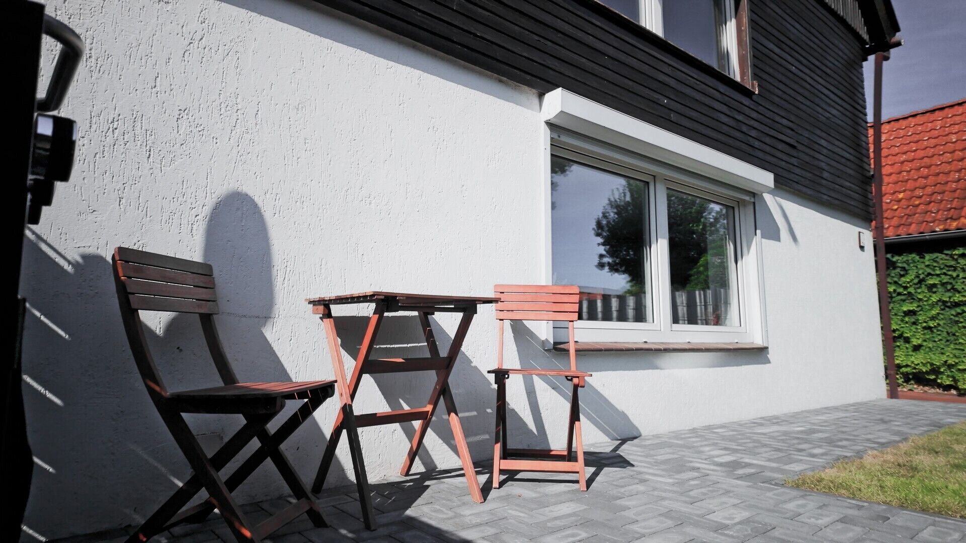 A patio with two wooden chairs and a table, a white wall, a window, and a red roof.