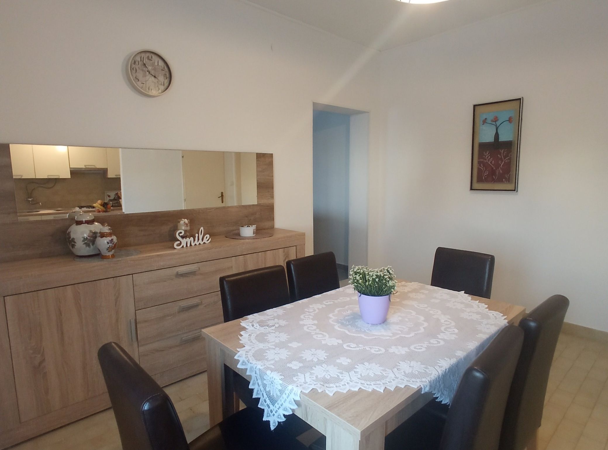 Un coin repas avec une table en bois, des chaises, une nappe en dentelle, une plante en pot et une horloge murale.
