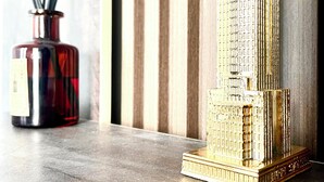Un modelo de un edificio alto con fachada detallada, un florero rojo con palitos negros y una pared texturizada de fondo.