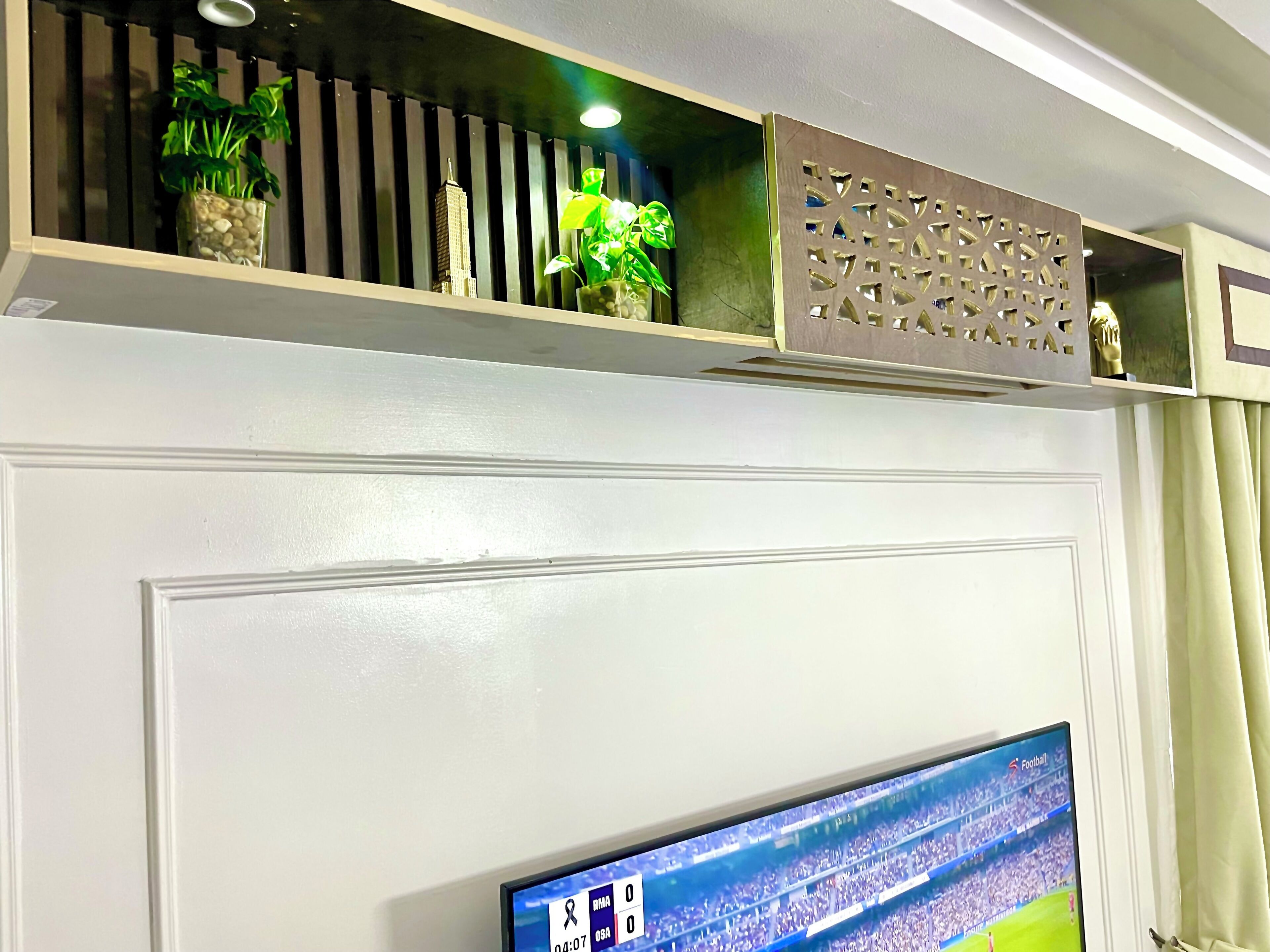 A living room with a flat-screen TV mounted on a white wall, a decorative shelf above it, and a curtain on the right.