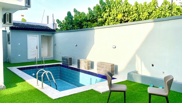 Una piscina con revestimiento de azulejos azules, rodeada de césped artificial verde. Hay dos sillas y una mesa pequeña cerca de la piscina.