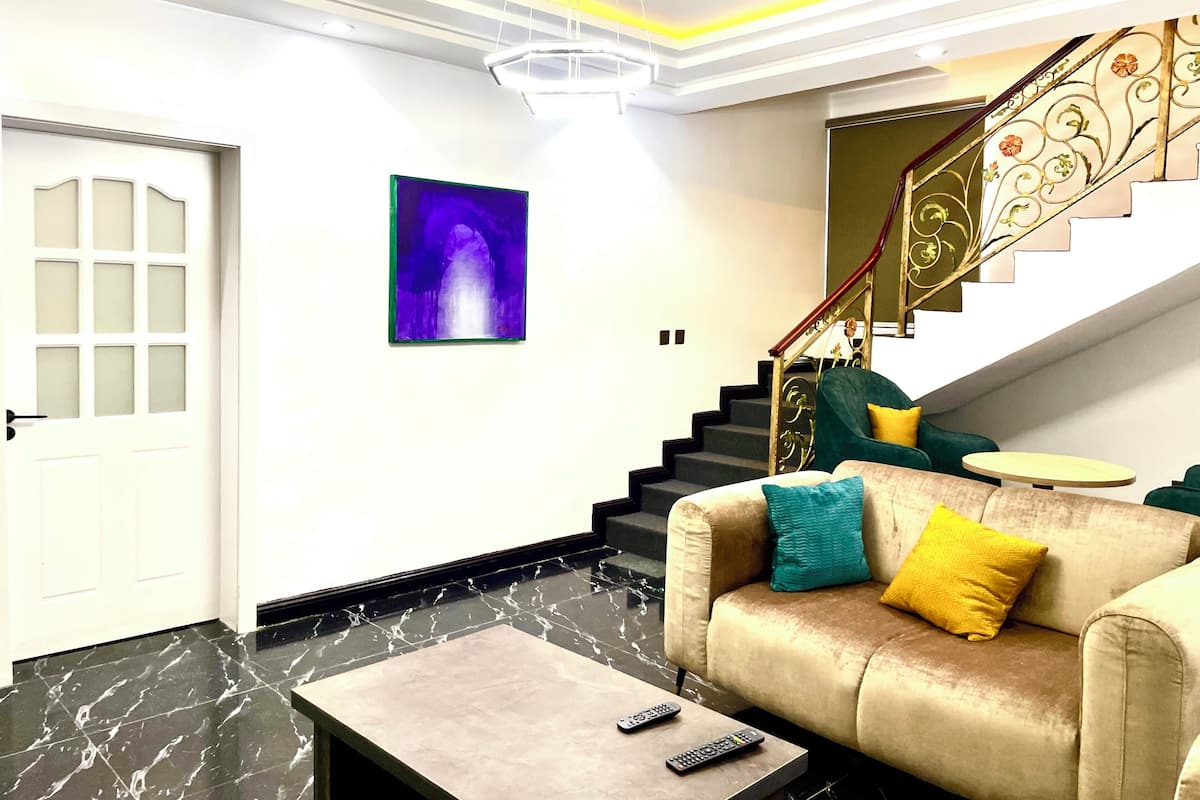 A modern living room with a marble floor, a beige sofa with colorful cushions, a coffee table, a painting on the wall, and a staircase with ornate railings.