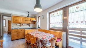 Een gezellige keuken met houten kasten, een eettafel met stoelen en grote ramen met gordijnen met een hartjespatroon.