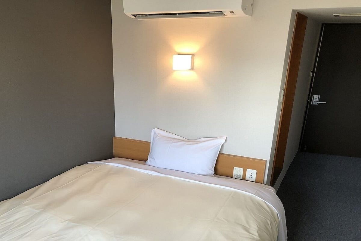 Una habitación de hotel con una cama, una lámpara fijada a la pared y una puerta.