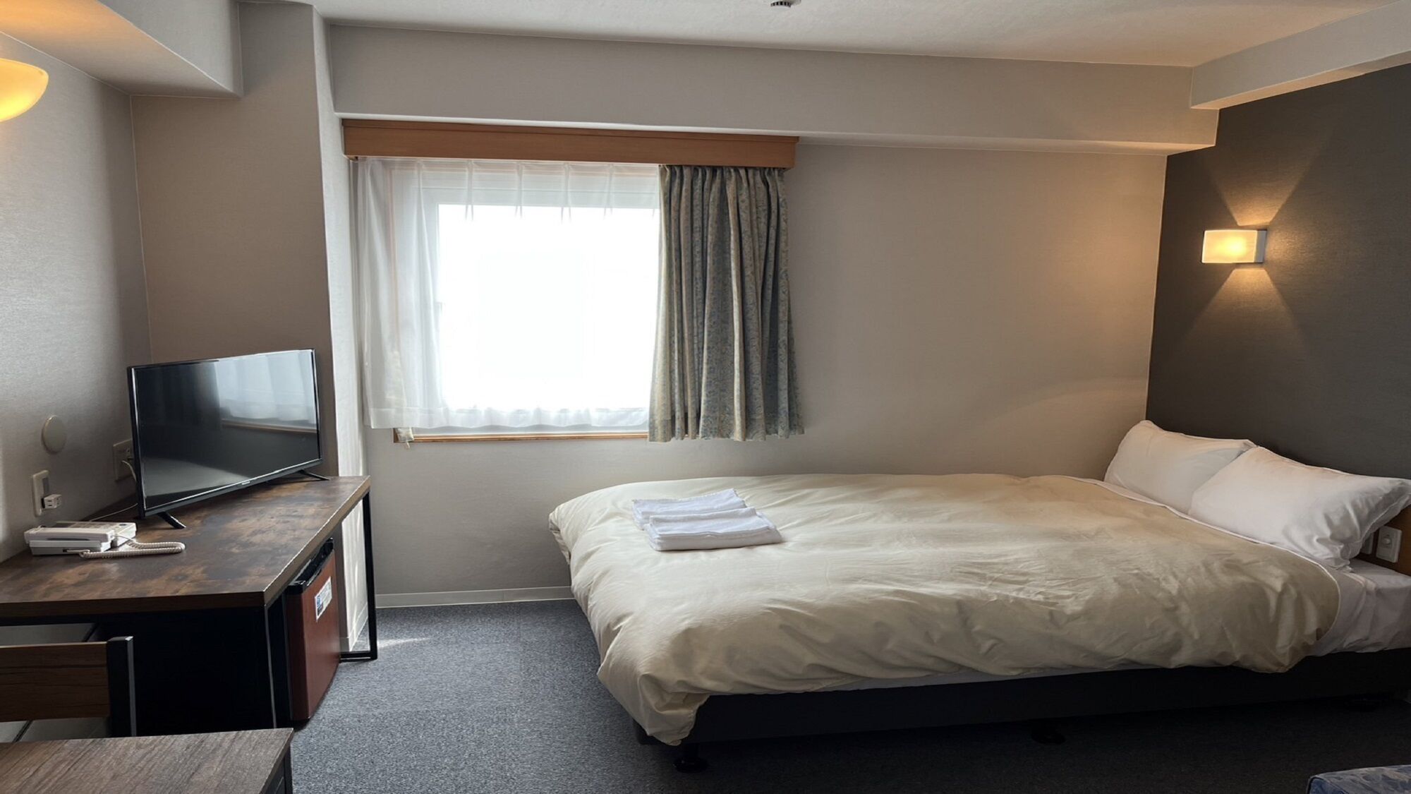 A hotel room with a bed, a desk with a TV, a window with curtains, and a wall-mounted light.